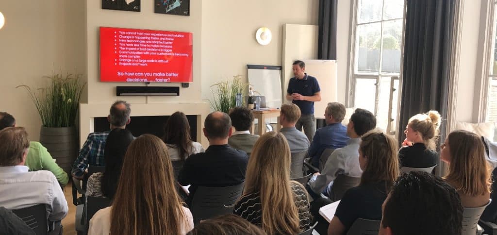 Spreker geeft presentatie voor zittend publiek bij een rode slide over betere en snellere besluiten